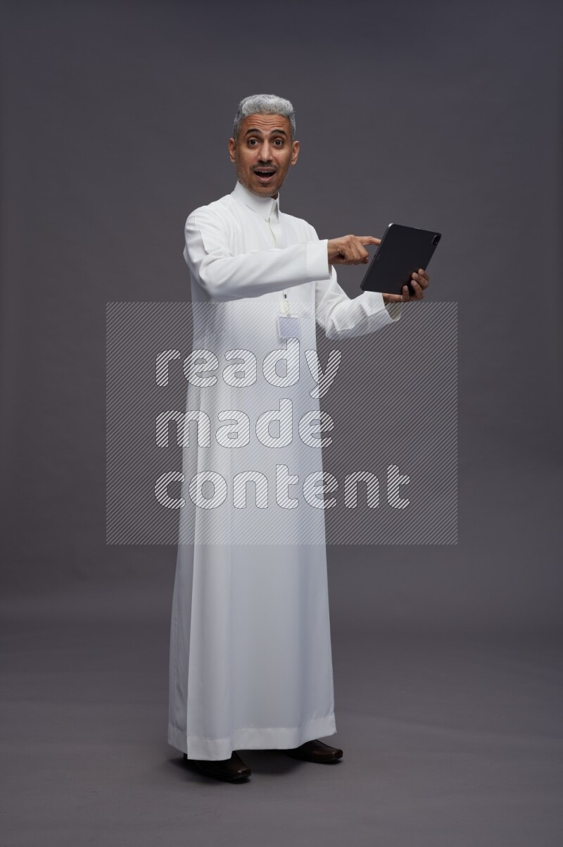 Saudi man wearing thob with neck strap employee badge standing working on tablet on gray background