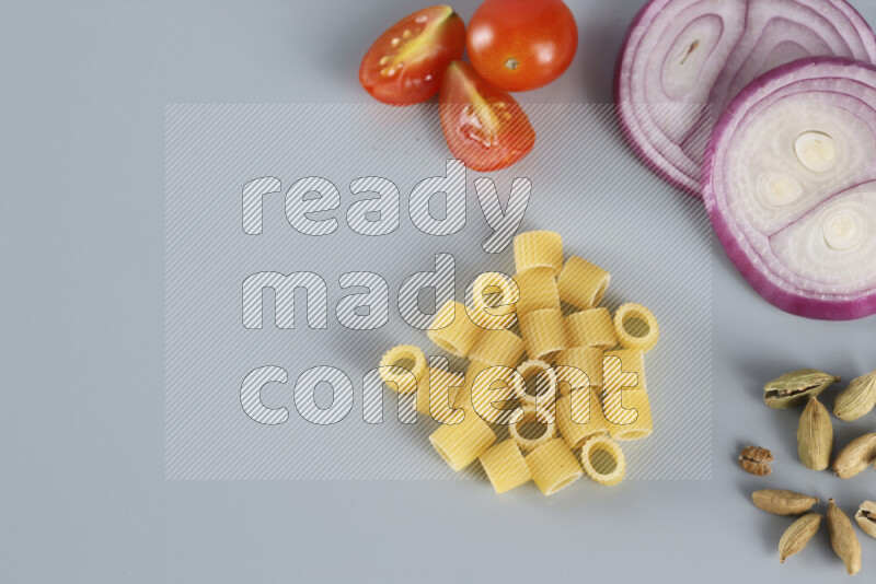 Raw pasta with different ingredients such as cherry tomatoes, garlic, onions, red chilis, black pepper, white pepper, bay laurel leaves, rosemary, cardamom and mushrooms on light blue background
