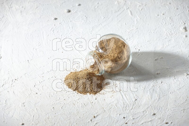 A flipped glass spice jar full of cumin powder and powder spilled out on textured white flooring