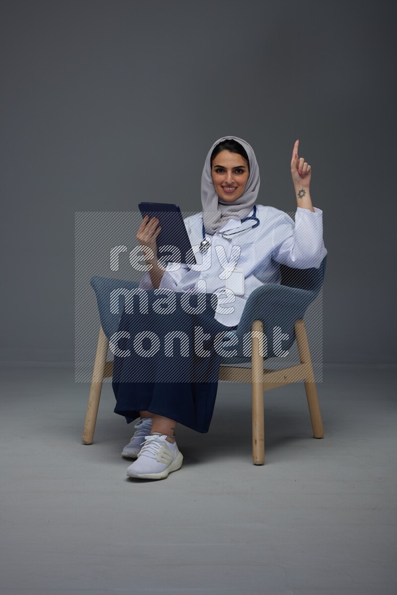 A doctor wearing a light gray head scarf setting on a dark grey chair and using a phone eye level on a grey background