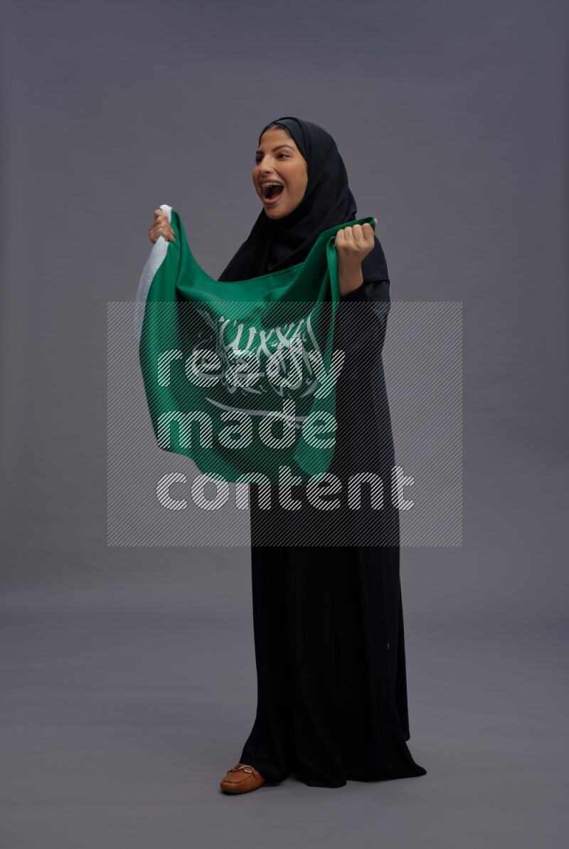 Saudi woman wearing Abaya standing holding Saudi flag on gray background