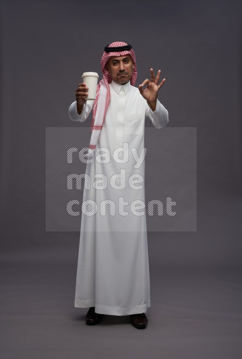 Saudi man wearing thob and shomag standing holding paper cup on gray background