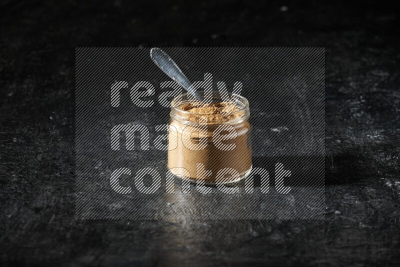 A glass jar and a metal spoon full of allspice powder on a textured black flooring