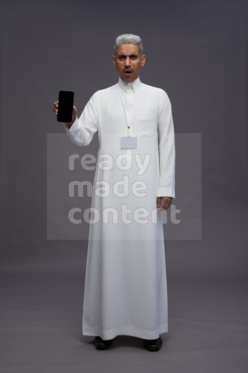 Saudi man wearing thob with neck strap employee badge standing showing phone to camera on gray background