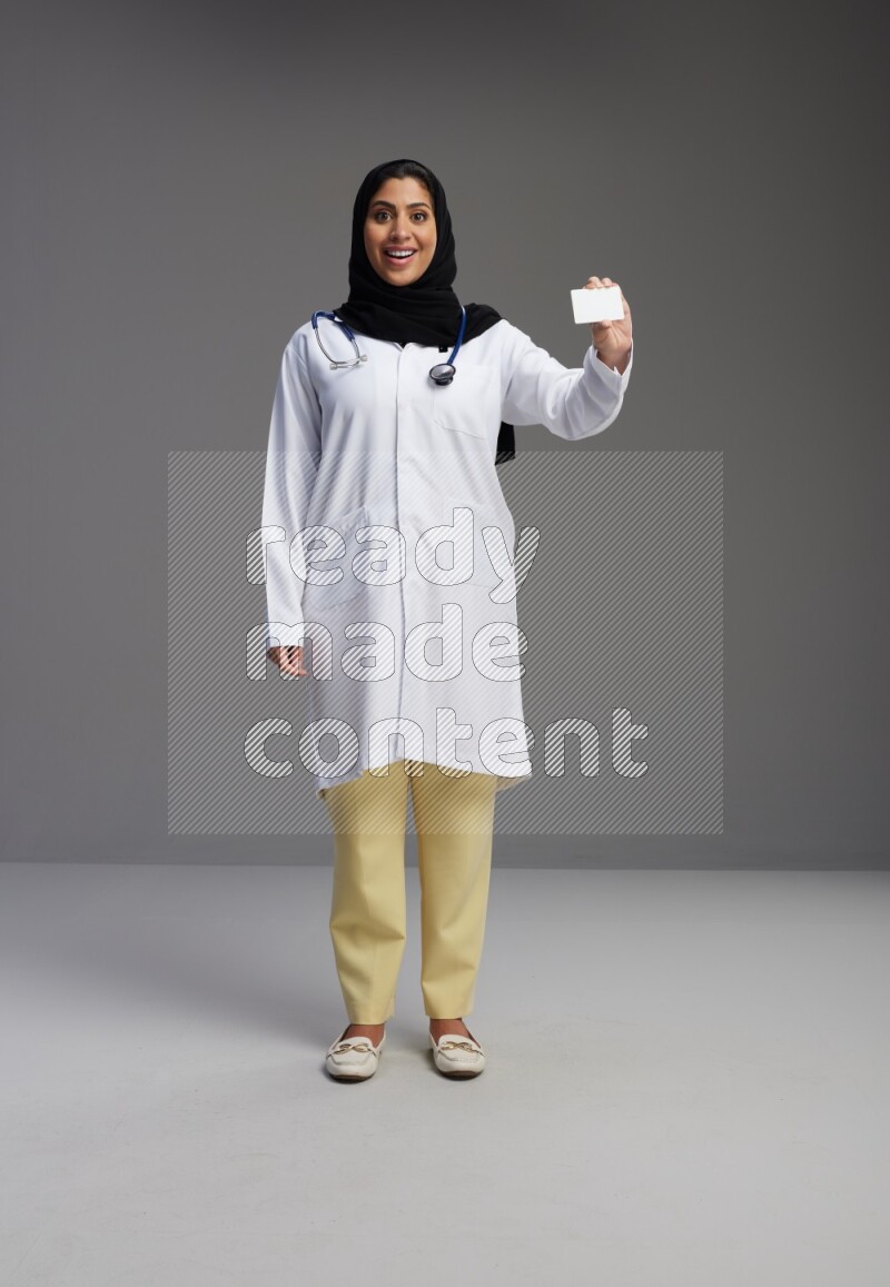 Saudi woman wearing lab coat with stethoscope standing holding ATM card on Gray background