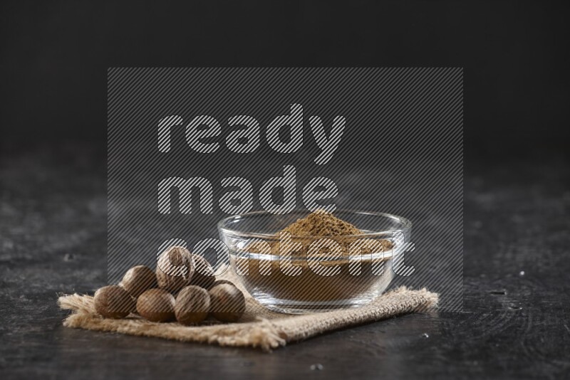 وعاء زجاجي ممتلئ ببودرة جوزة الطيب مع بعض الحبوب بجانبه على قماش الخيش على خلفية سوداء