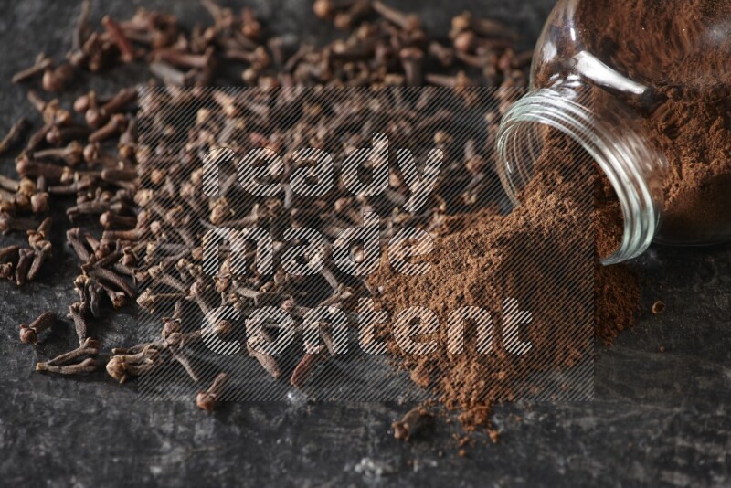 وعاء زجاجي للبهارات مقلوب ممتلئ ببودرة القرنفل والبودرة سقطت منه مع حبوب القرنفل المتناثرة على أرضية سوداء