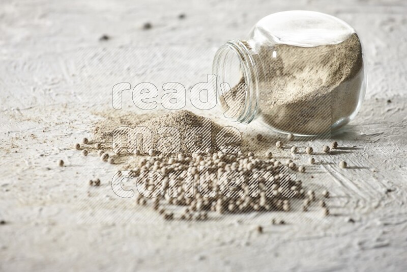 A flipped herbal glass jar full of white pepper powder with spilled powder and beads on textured white flooring