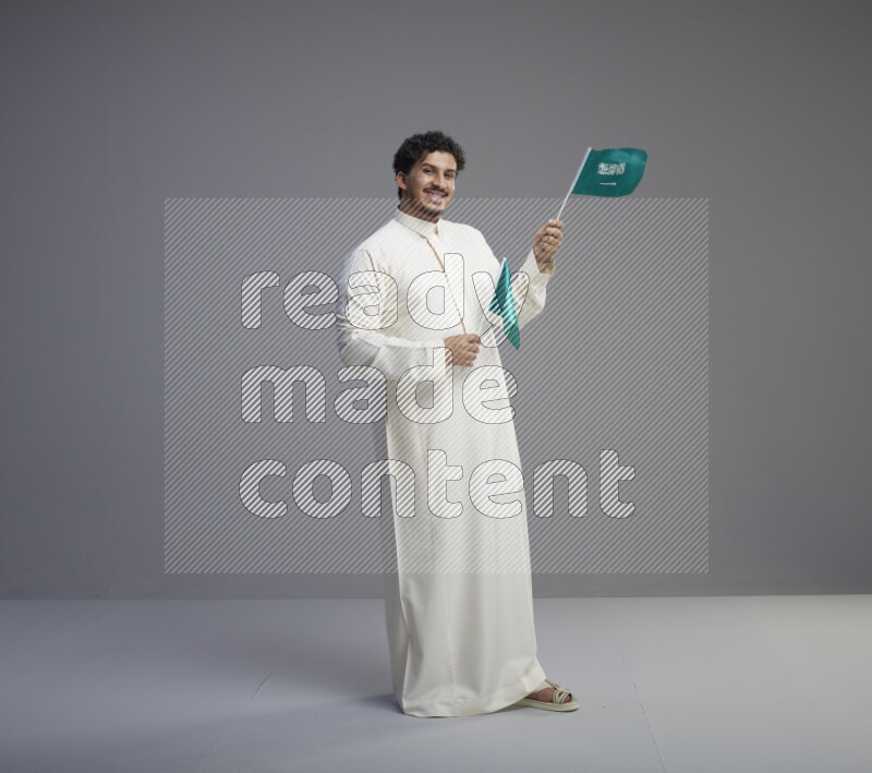 A Saudi man standing wearing thob holding small Saudi flag on gray background