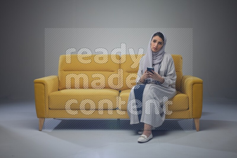 A Saudi woman wearing a light gray Abaya and white head scarf sitting on a yellow sofa and using her phone eye level on a grey background