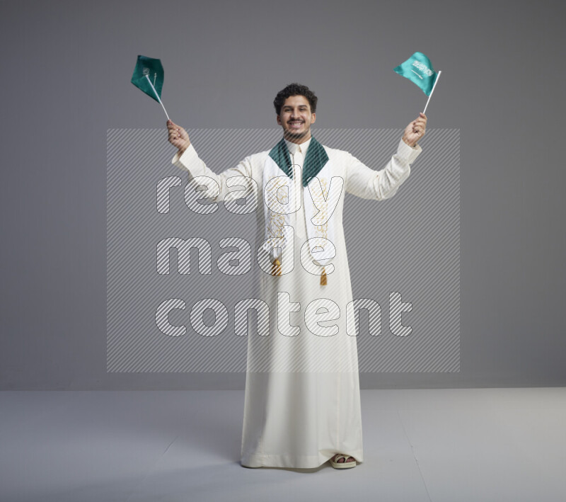 A Saudi man standing wearing thob and saudi flag scarf and holding small saudi flag on gray background