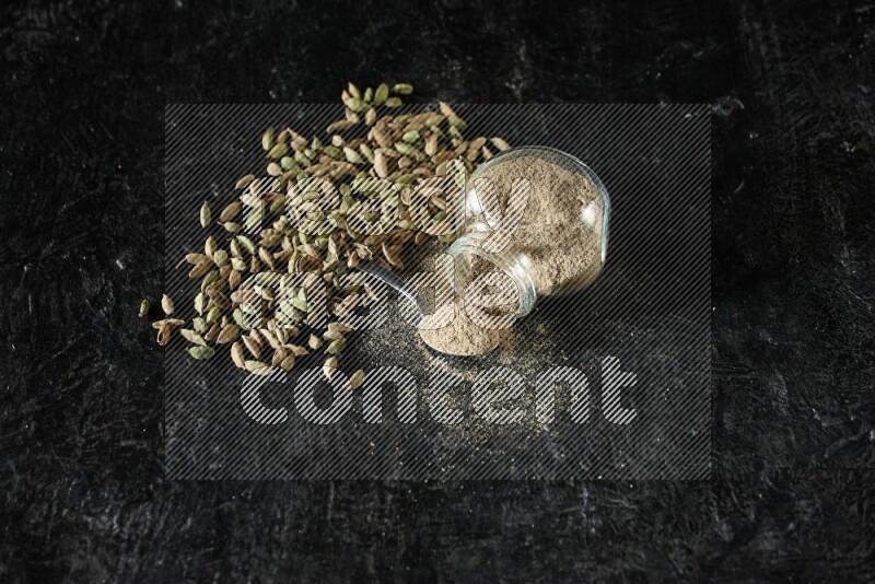 وعاء زجاجي للتوابل مقلوب وملعقة معدنية ممتلئة ببودرة الهيل وحبوب الهيل متناثرة على خلفية سوداء