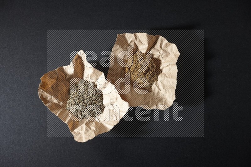 Cumin seeds and powder in 2 crumpled pieces of paper on black flooring