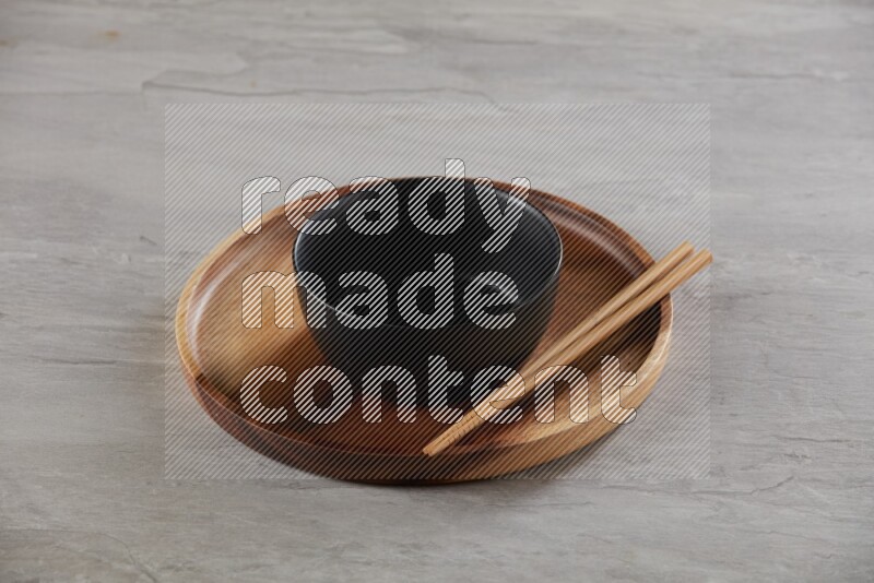 black ceramic round bowl on top of brown wood round plate and wood chopsticks, on grey textured countertop