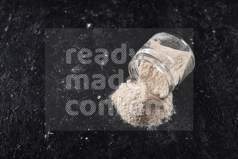 A glass jar full of onion powder flipped with some spilling powder on black background