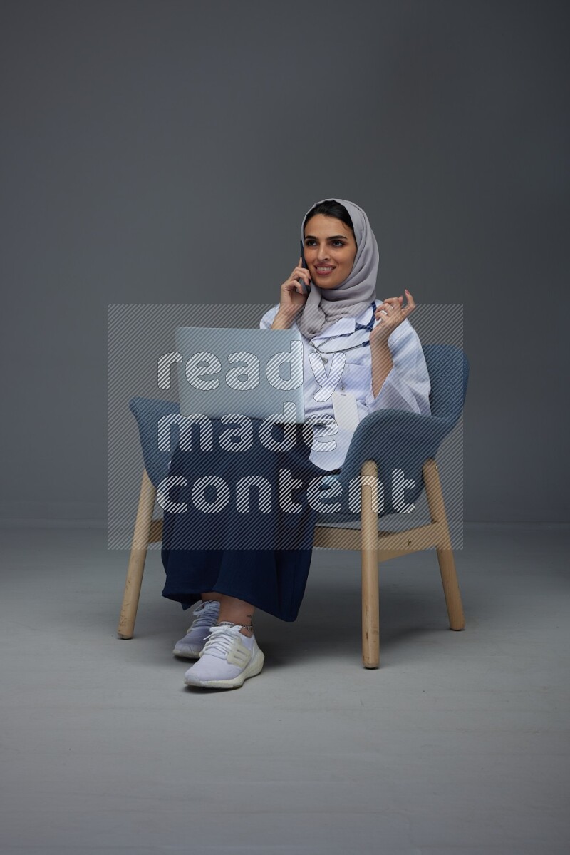 A doctor wearing a light gray head scarf sitting on blue chair on grey background