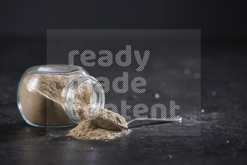 A glass spice jar full of garlic powder flipped and the powder came out with a filled metal spoon beside it on a textured black flooring
