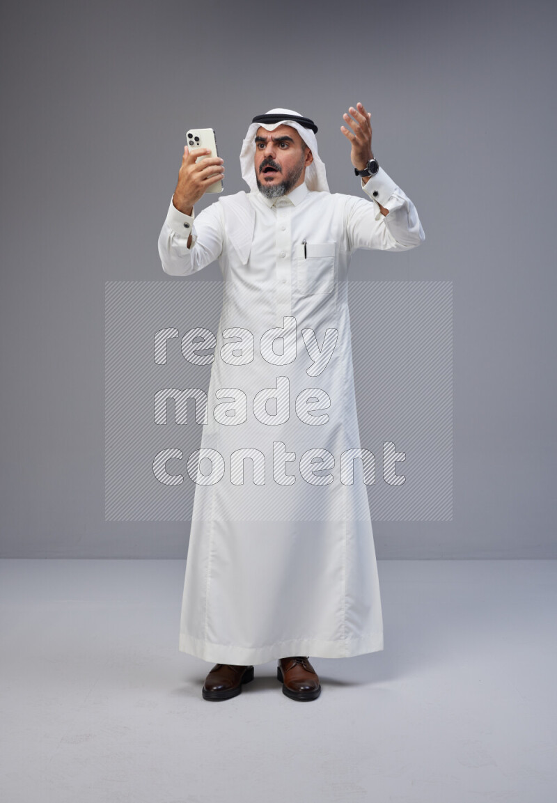 Saudi man Wearing Thob and white Shomag standing texting on phone on Gray background