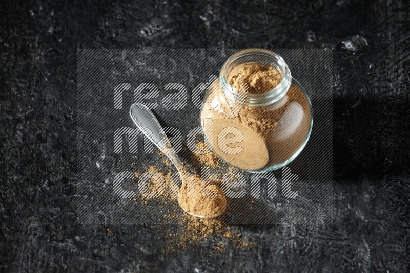 A glass spice jar and metal spoon full of allspice powder on a textured black flooring