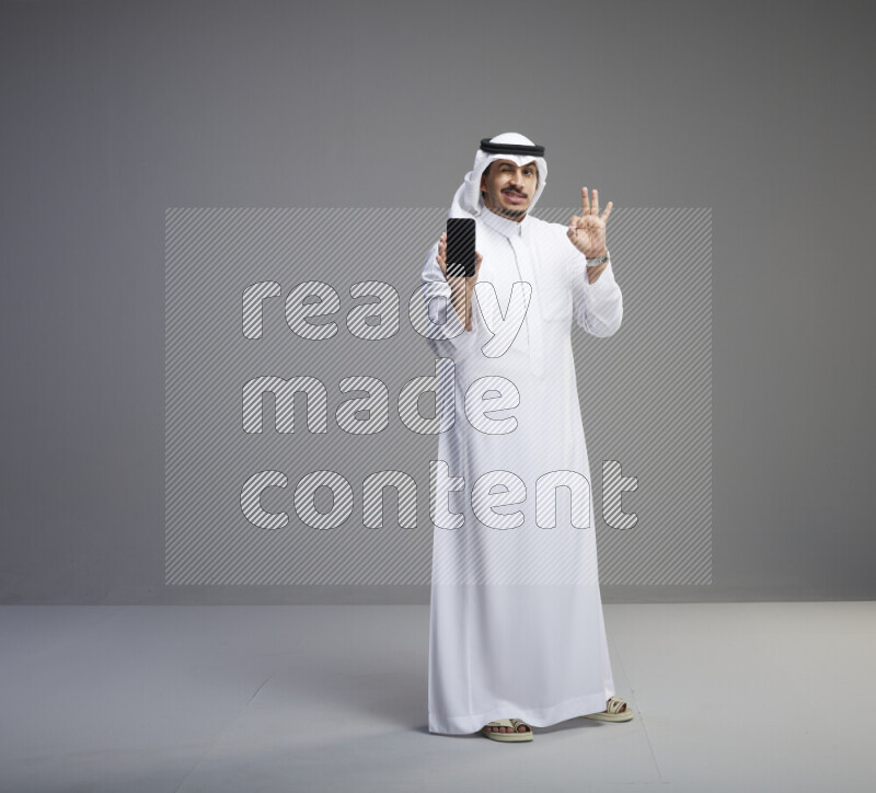 A Saudi man standing wearing thob and white shomag showing phone to camera on gray background