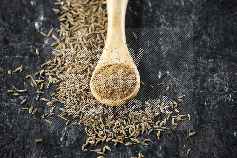 A wooden spoon full of cumin powder and cumin seeds spreaded on textured black flooring
