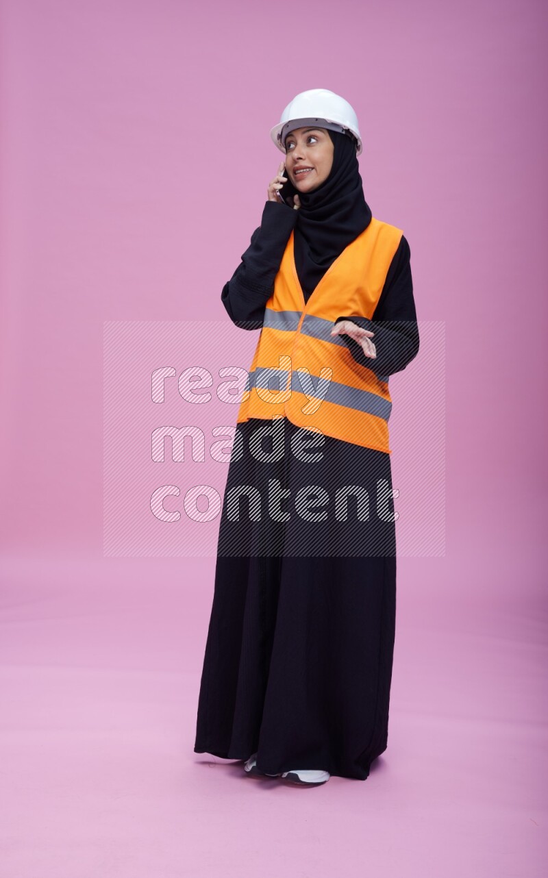 Saudi woman wearing Abaya with engineer vest and helmet standing talking on phone on pink background