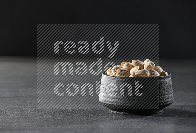 A black pottery bowl full of pistachios on a black background in different angles