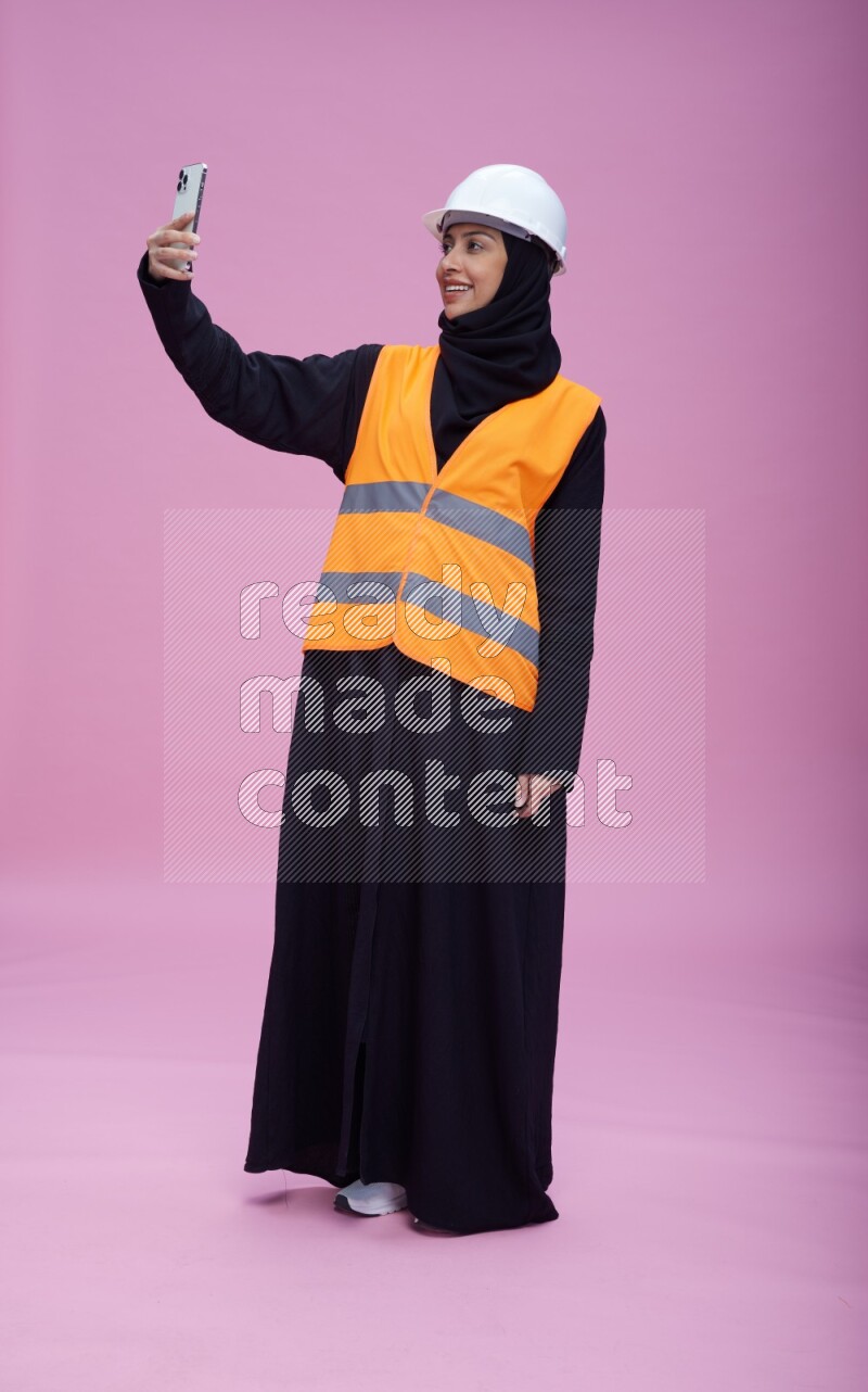 Saudi woman wearing Abaya with engineer vest and helmet standing taking selfie on pink background