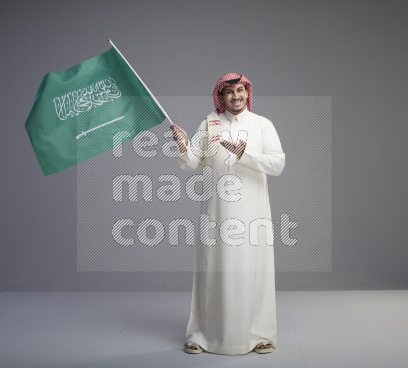 A Saudi man standing wearing thob and red shomag raising big Saudi flag on gray background