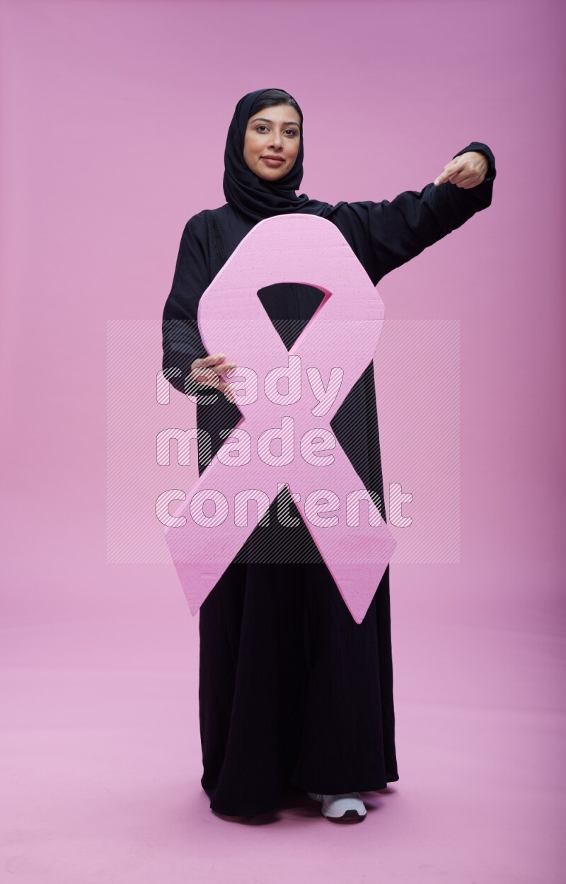 Saudi woman wearing pink ribbon on abaya standing holding awareness ribbon on plain pink background