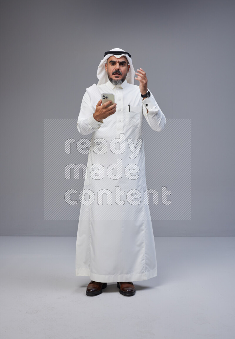 Saudi man Wearing Thob and white Shomag standing texting on phone on Gray background