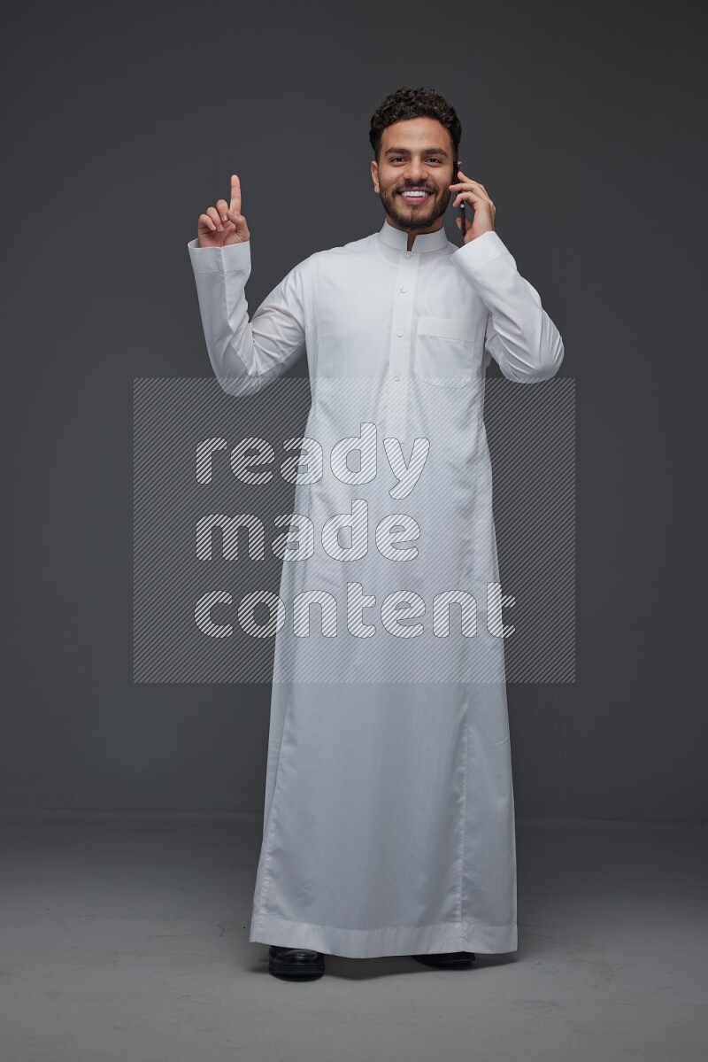 A Saudi man wearing Thobe and talking in the phone while standing and making different hand gestures eye level on a gray background