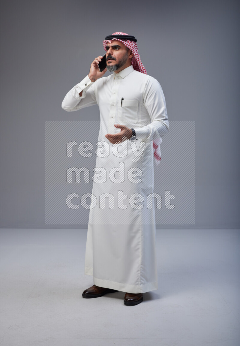 Saudi man Wearing Thob and red Shomag standing talking on phone on Gray background