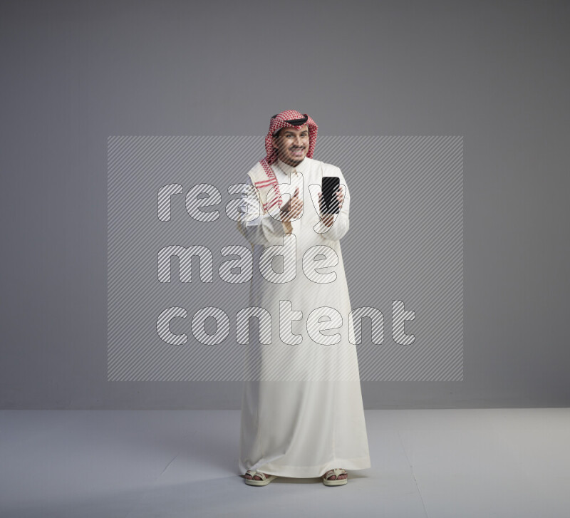 A Saudi man standing wearing thob and red shomag showing phone to camera on gray background