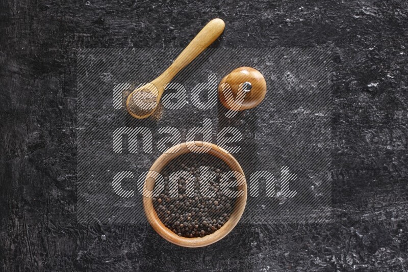 A wooden bowl full of black pepper and a wooden spoon full of black pepper powder and a wooden grinder on a textured black flooring