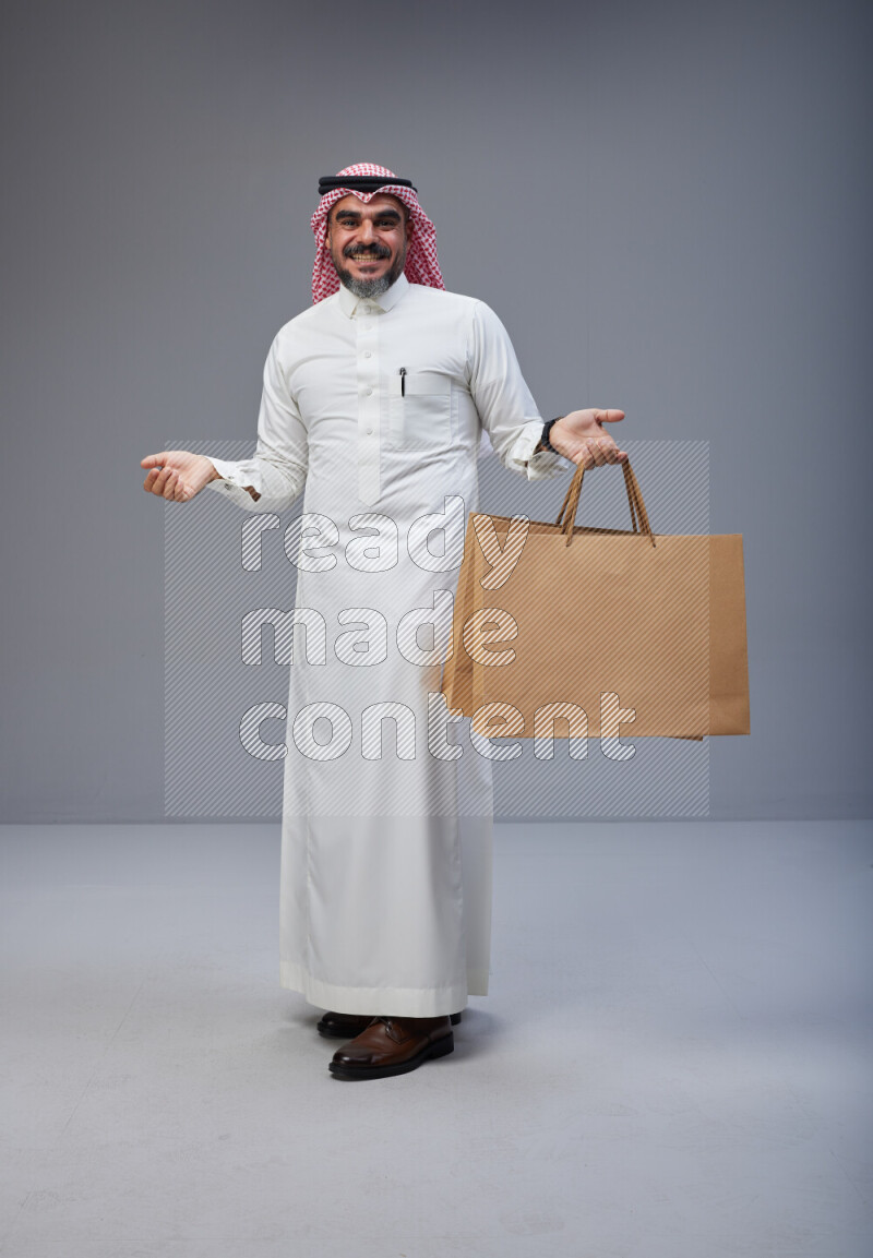 Saudi man Wearing Thob and red Shomag standing holding shopping bag on Gray background