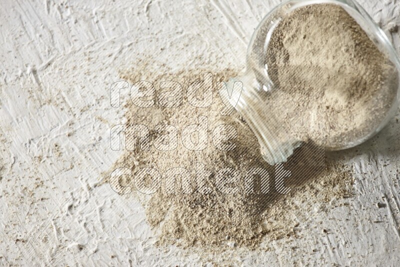 A flipped herbal glass jar full of white pepper powder with spilled powder on textured white flooring