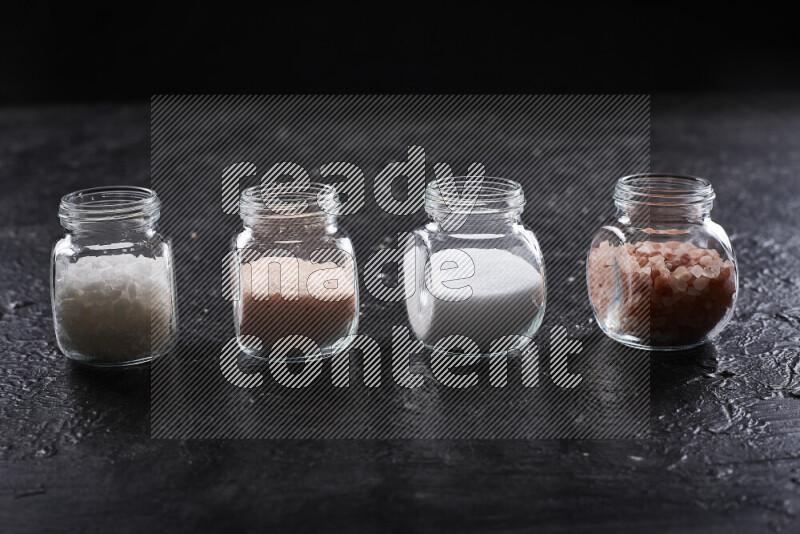 4 glass jars filled with table salt, coarse sea salt, fine himalayan salt and coarse himalayan salt on black background