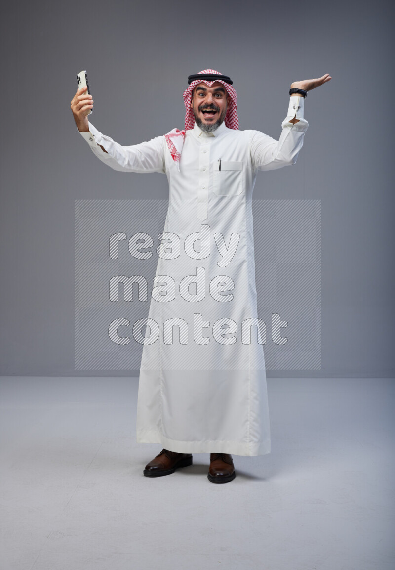 Saudi man Wearing Thob and red Shomag standing taking selfie on Gray background