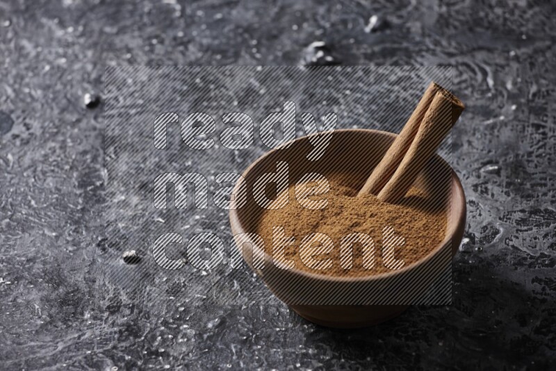 Wooden bowl full of cinnamon powder and a cinnamon stick on a textured black background