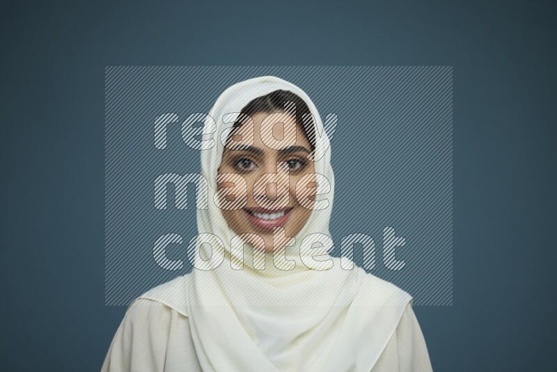 A Saudi woman posing in a blue background wearing an off-white Abaya with hijab