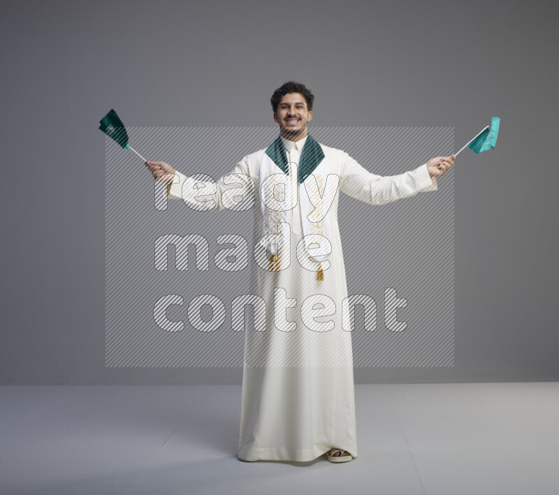 A Saudi man standing wearing thob and saudi flag scarf and holding small saudi flag on gray background