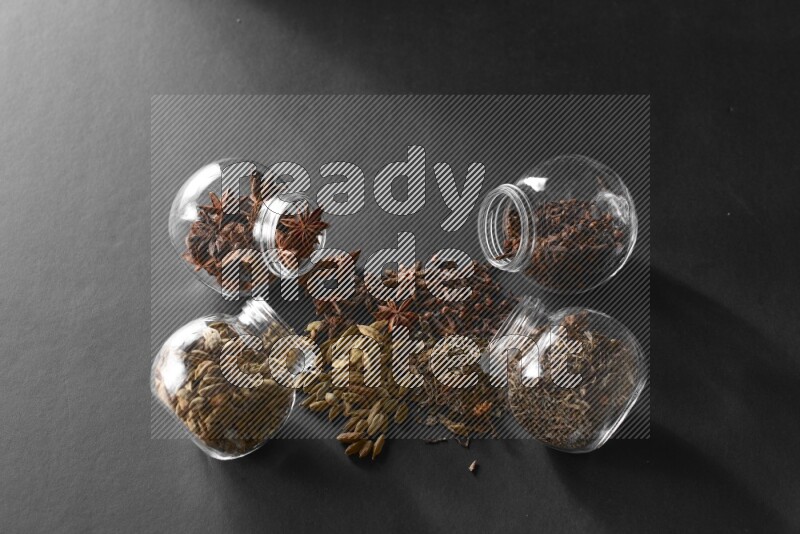4 glass spice jars full of cloves, cardamom, basil and star anise on black flooring