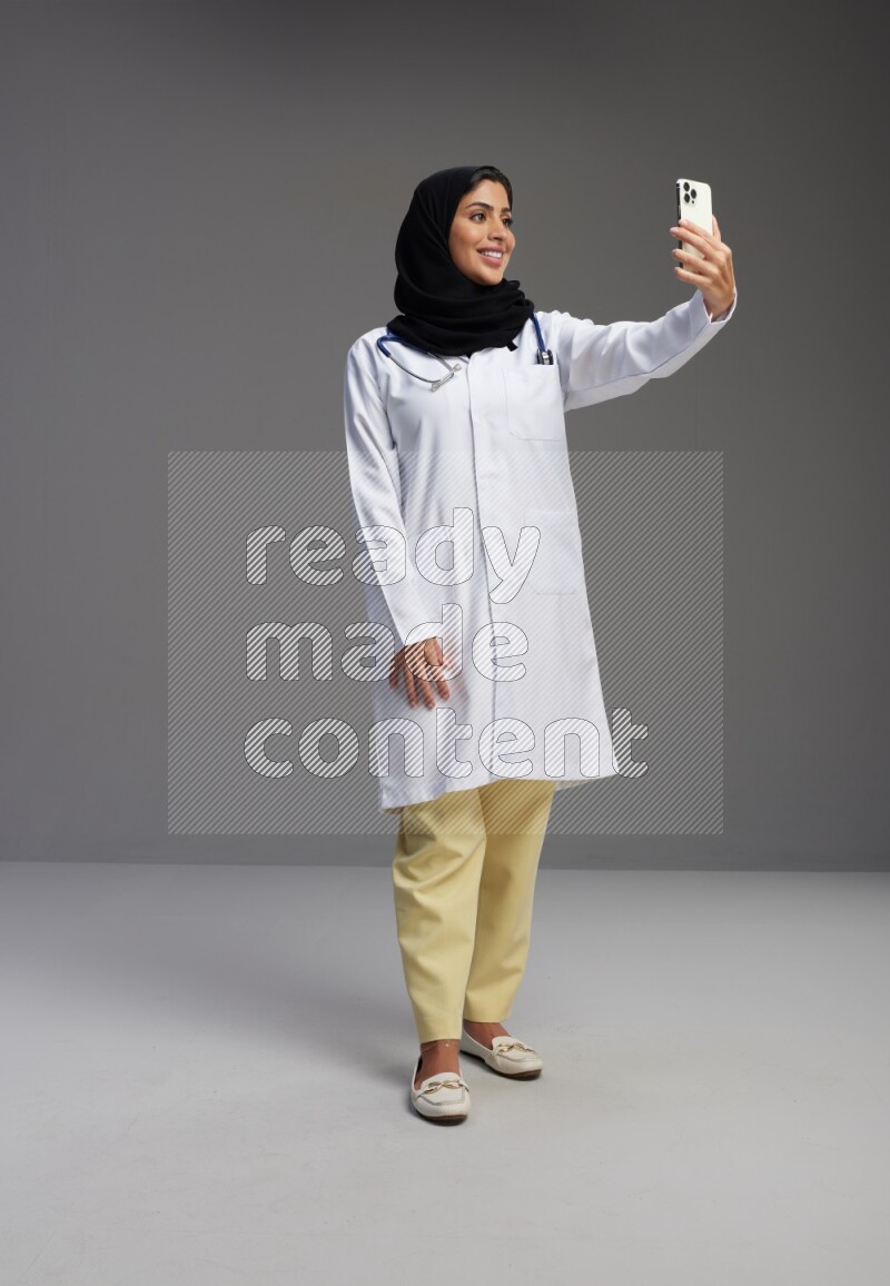 Saudi woman wearing lab coat with stethoscope standing taking selfie on Gray background