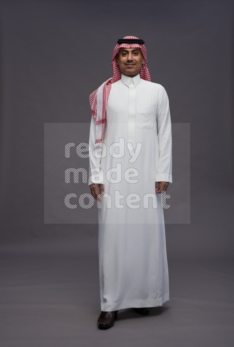 Saudi man wearing thob and shomag standing interacting with the camera on gray background