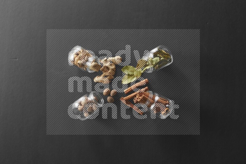 4 glass spice jars full of laurel bay leaves, ginger, cinnamon and nutmeg on black flooring