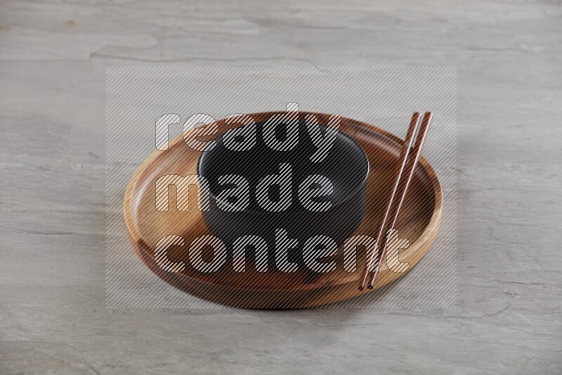 black ceramic round bowl on top of brown wood round plate and wood chopsticks, on grey textured countertop