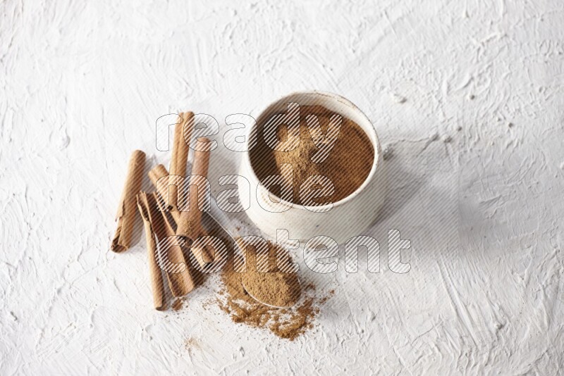 Ceramic beige bowl full of cinnamon powder and a metal spoon with cinnamon sticks next of it on a textured white background