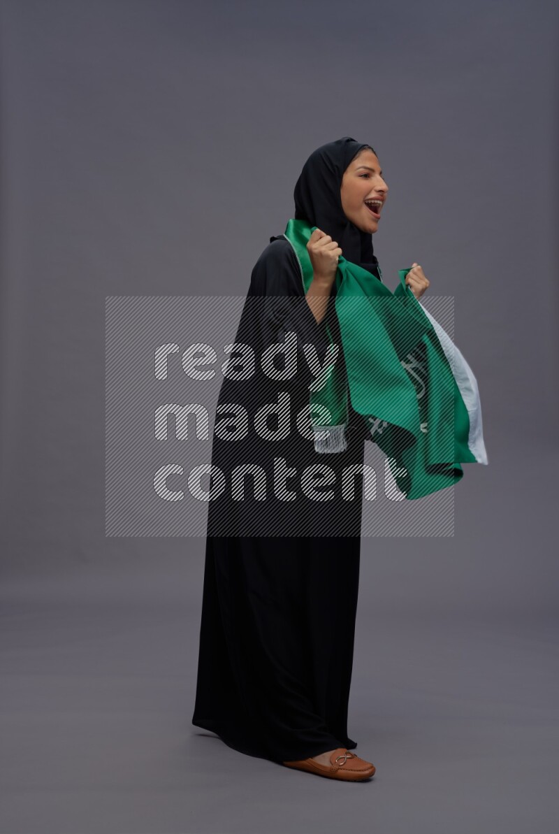 Saudi woman wearing Abaya standing holding Saudi flag on gray background