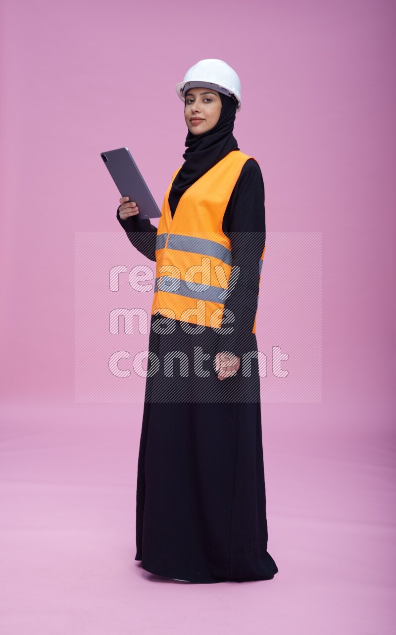 Saudi woman wearing Abaya with engineer vest and helmet standing working on tablet on pink background